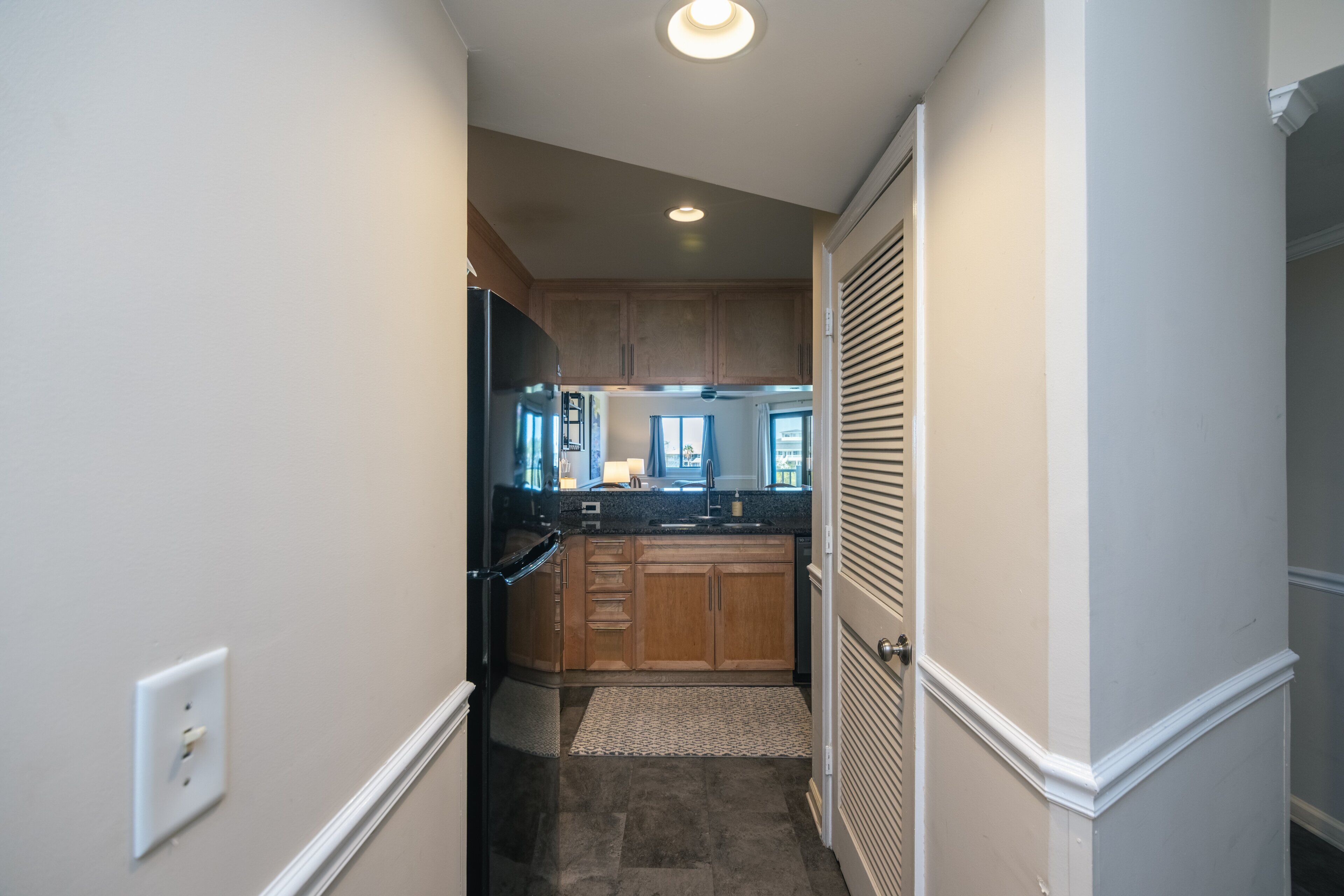 A narrow hallway leading to a kitchen with wooden cabinets, a black refrigerator, and a window with curtains.