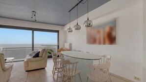 A modern living room with a large window, a round dining table, and a sofa. - Sea view apartment at the pier. (Blankenberge)