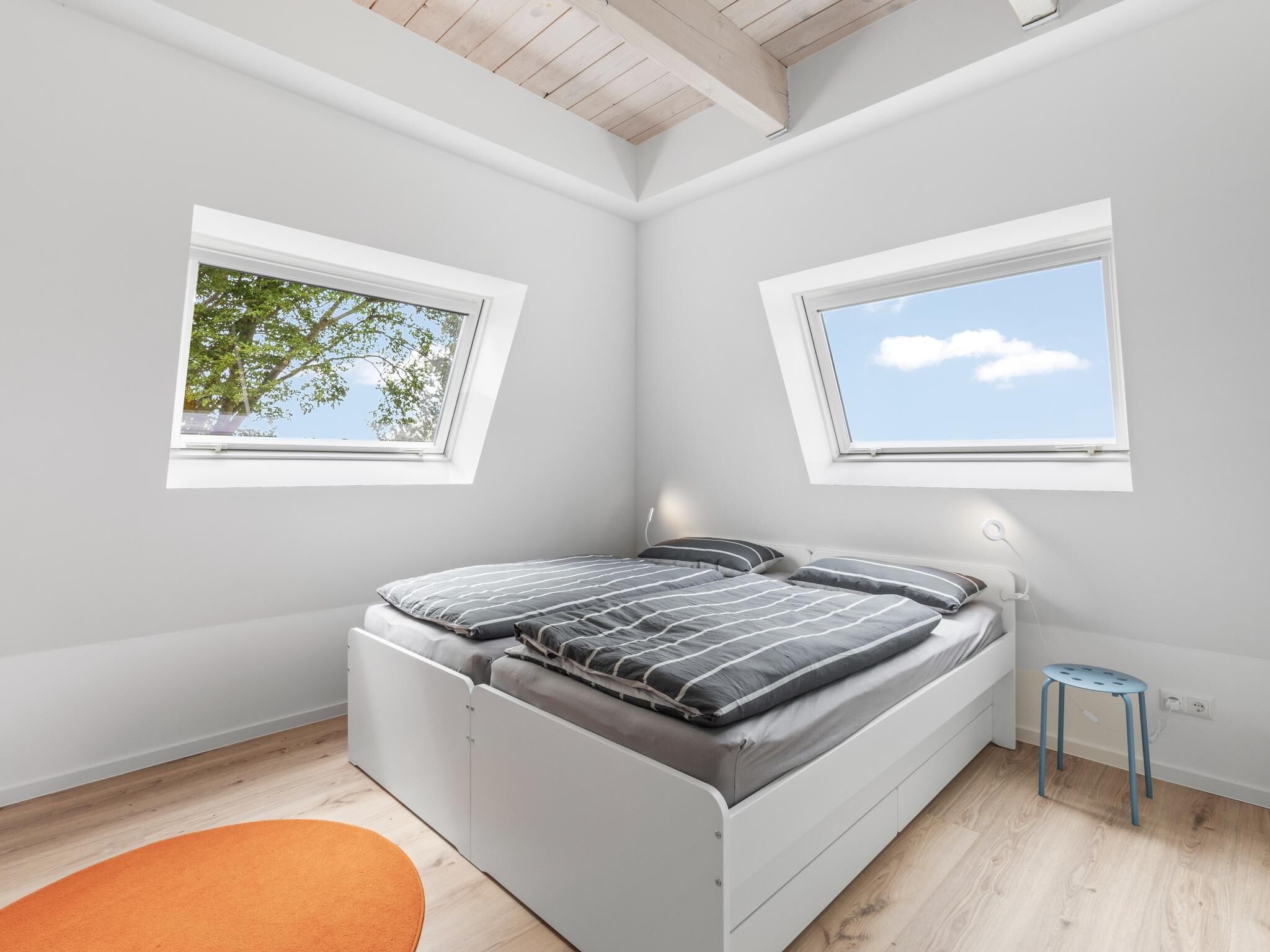 A bedroom with a bed, two windows, a small stool, and a wooden ceiling.