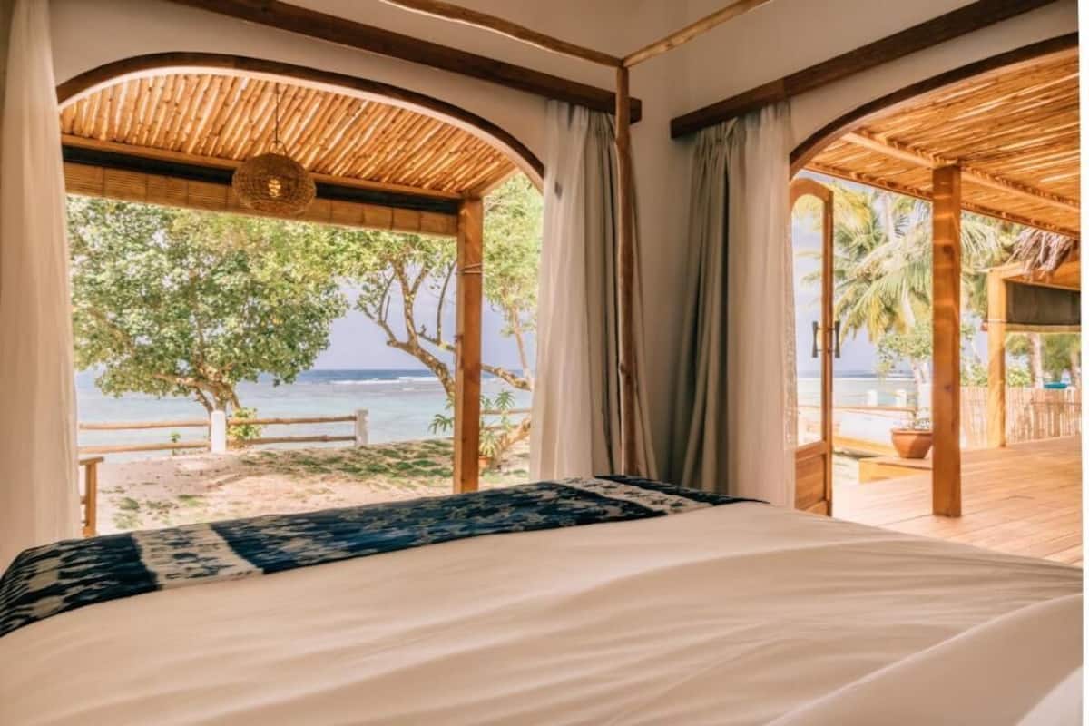 A bedroom with a bed, a view of the beach, and a wooden ceiling structure.