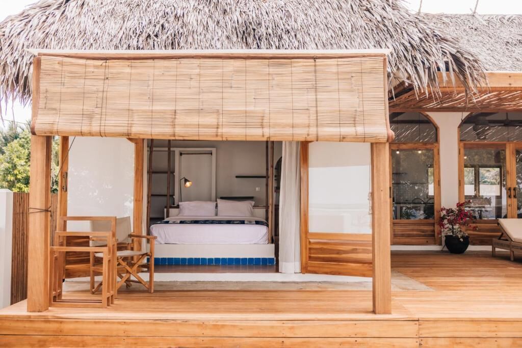 Una terraza de madera con techo de paja, una cama con ropa blanca y una pequeña mesa con una silla.
