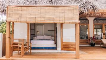 Una terraza de madera con techo de paja, una cama con ropa blanca y una pequeña mesa con una silla.