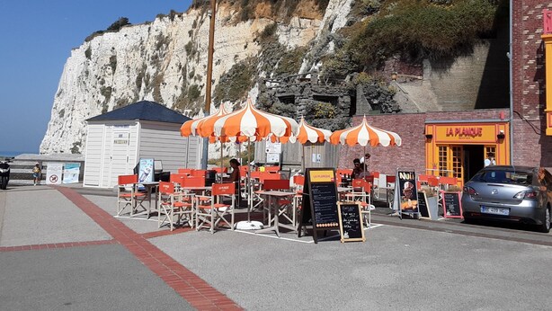 A seaside café with outdoor seating, a cliffside backdrop, and a building with a red and yellow facade.