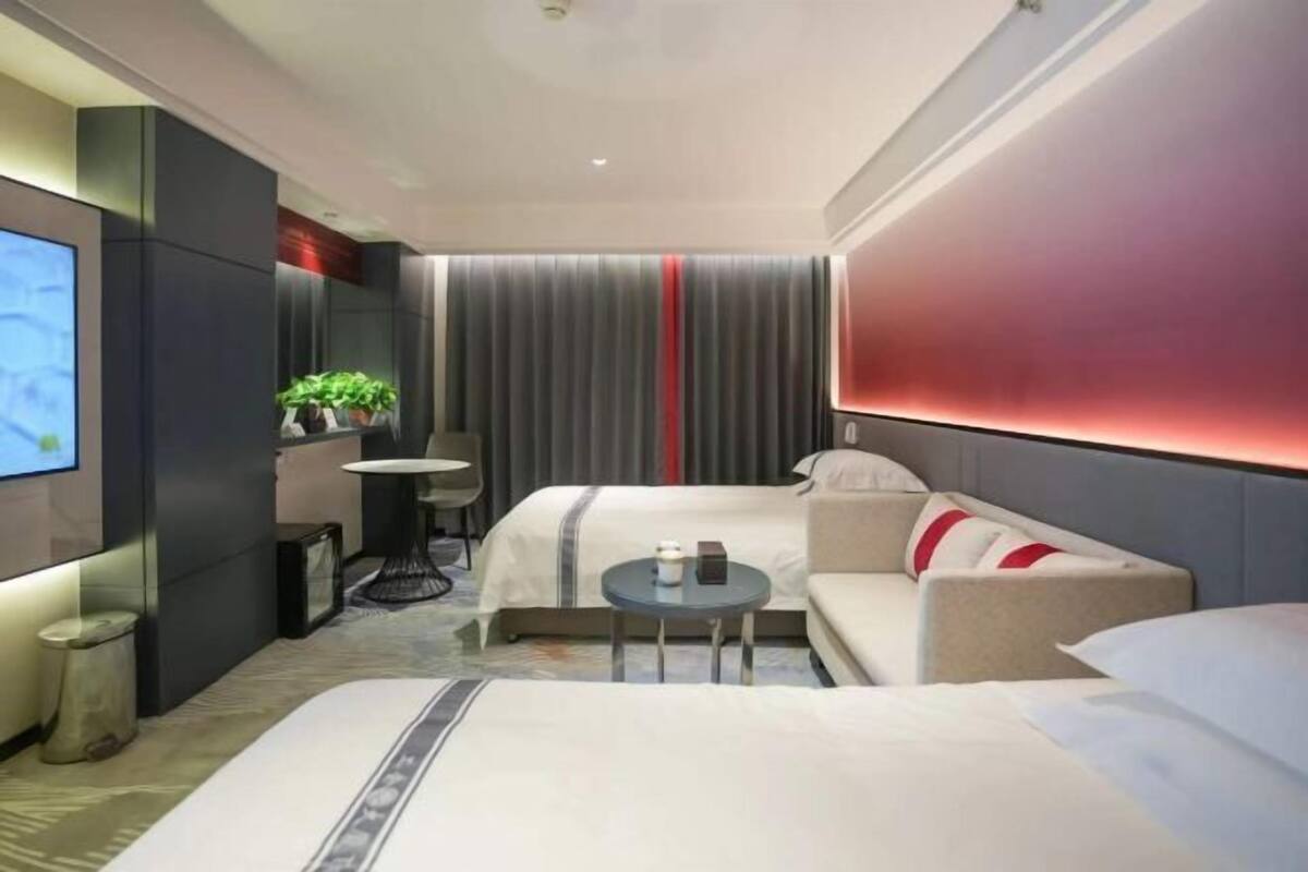 A modern hotel room with a bed, a sofa, a small table, a TV, and a plant.