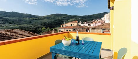 A balcony with a table, chairs, a bowl of fruit, and a bottle of wine.