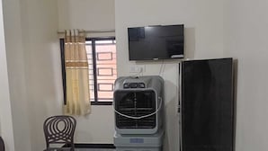 Una habitación con televisor montado en la pared, un refrigerador, una silla y una mesa con mantel floral.