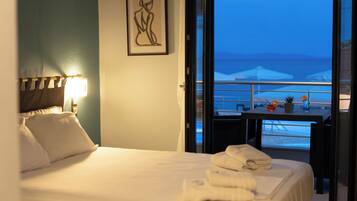 A hotel room with a bed, a painting on the wall, a balcony with a view of the sea, and a table with a plant and a vase.