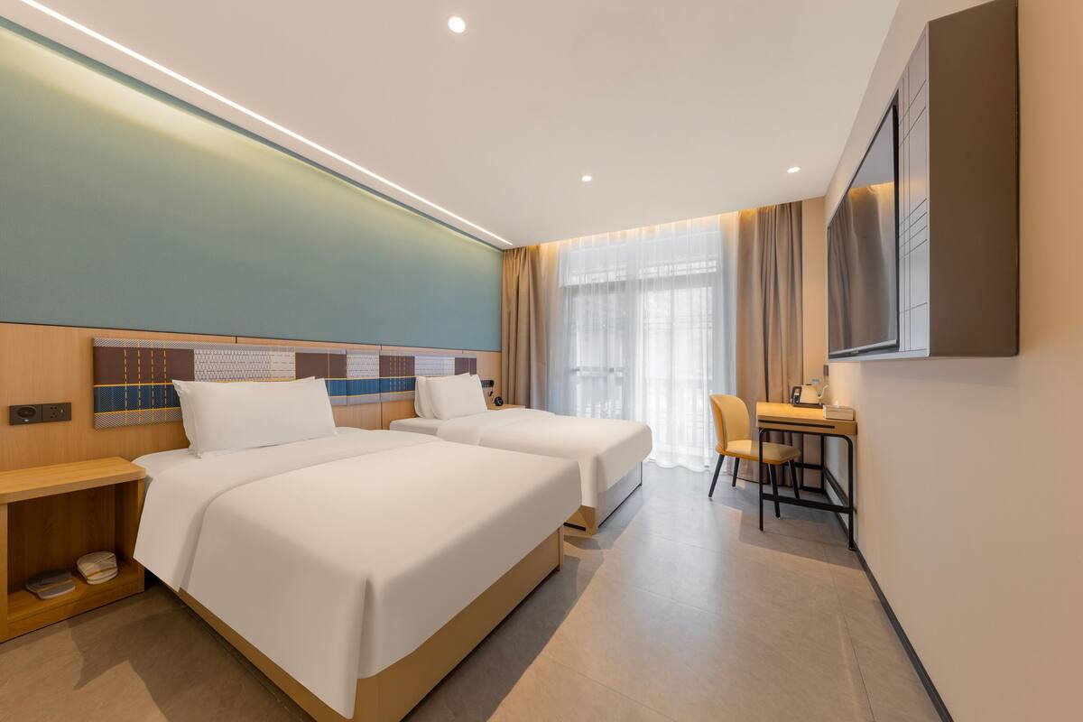 Ein Hotelzimmer mit zwei Betten, einem Schreibtisch, einem Stuhl, einem Fernseher und einem Fenster mit Vorhängen.