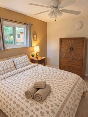 A bedroom with a bed, a nightstand, a lamp, a ceiling fan, a window, and a wooden cabinet.