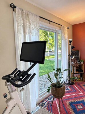 A stationary bike with a screen, a window with curtains, a plant, and a welcome sign.
