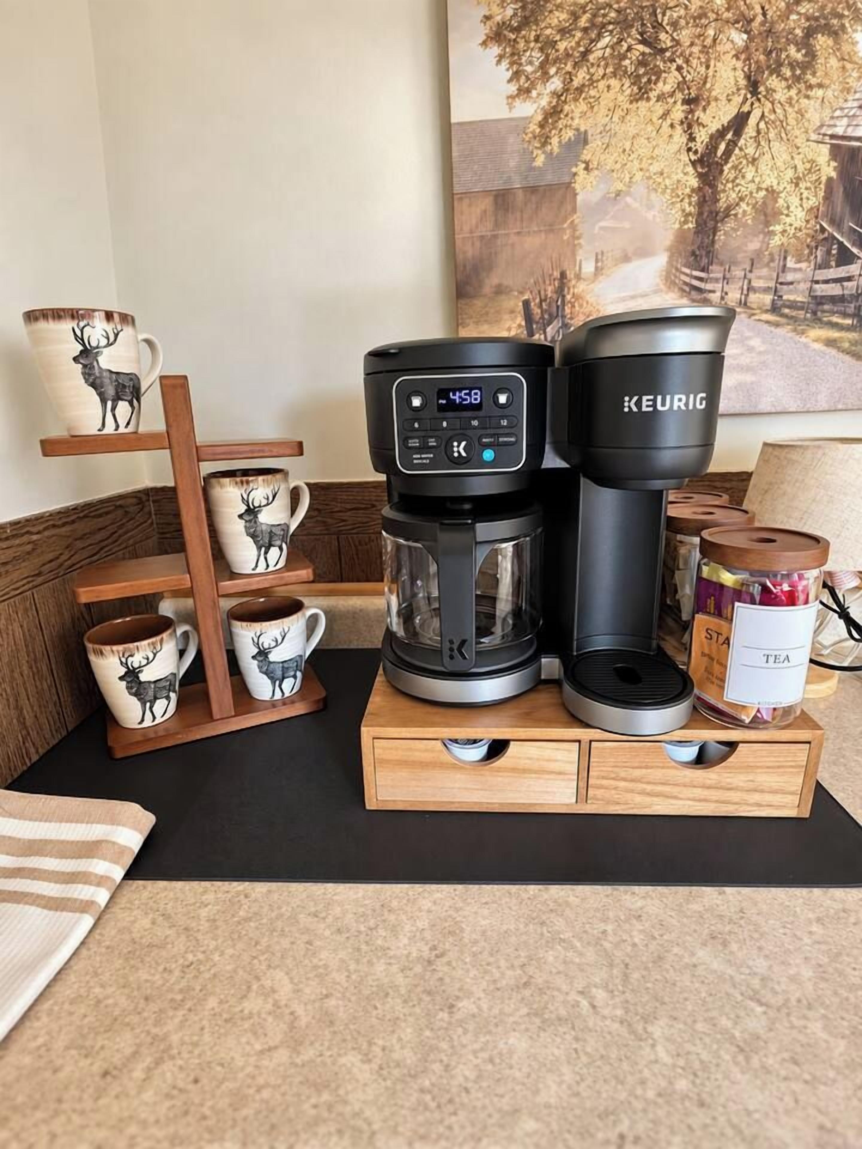 Eine Kaffeemaschine auf einem Holzständer mit einer Tassenhalterung, einer mit 'TEE' beschrifteten Dose und einem Gemälde im Hintergrund.