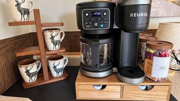 Eine Kaffeemaschine auf einem Holzständer mit einer Tassenhalterung, einer mit 'TEE' beschrifteten Dose und einem Gemälde im Hintergrund.
