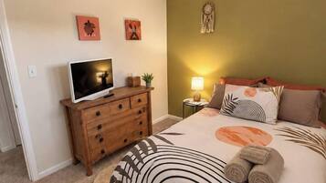 Quarto com cama, ventoinha no teto, televisão sobre cômoda de madeira e detalhes decorativos nas paredes.