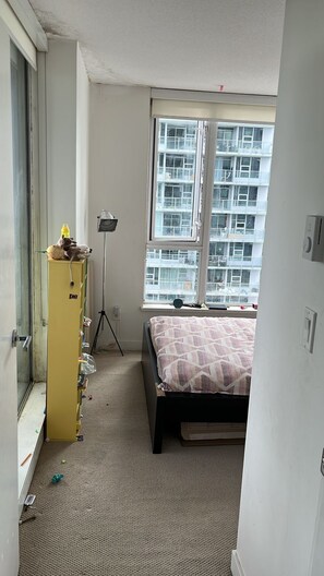 A bedroom with a bed, a window with a view of a building, a yellow cabinet, and a tripod lamp.