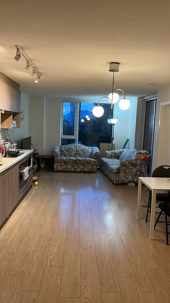 A modern kitchen with wooden flooring, a dining table, two floral sofas, and a large window with a view of mountains.