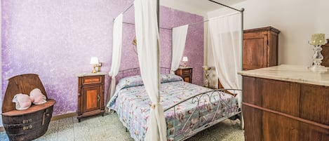 A four-poster bed with a floral bedspread, a wooden nightstand with a lamp, a vintage chair, and a wooden cabinet.