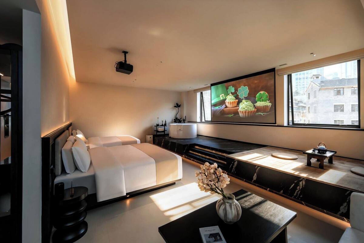 A modern hotel room with a bed, a desk, a large window, and a painting of cupcakes.