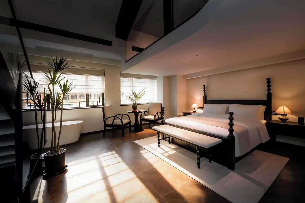 A modern hotel room with a large bed, a seating area with chairs, a potted plant, and a view of the city through large windows.
