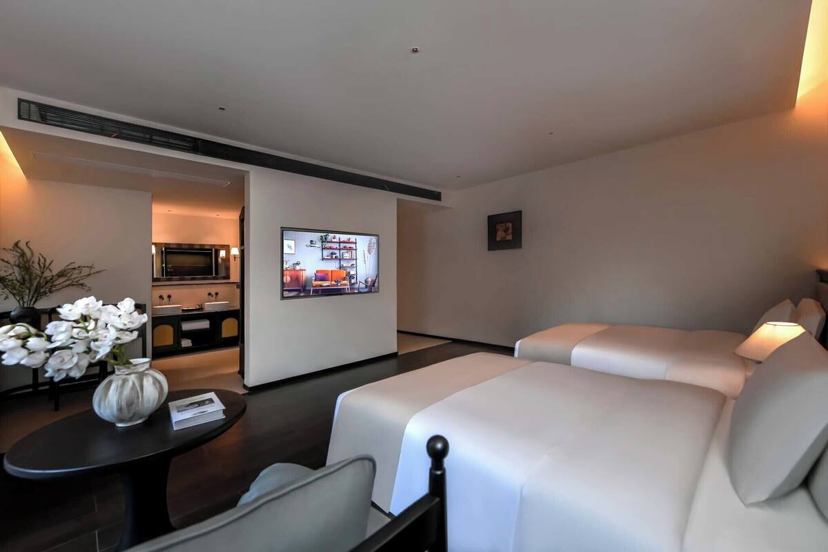 A hotel room with a TV, two beds, a small table with a vase, and a view into the kitchen area.