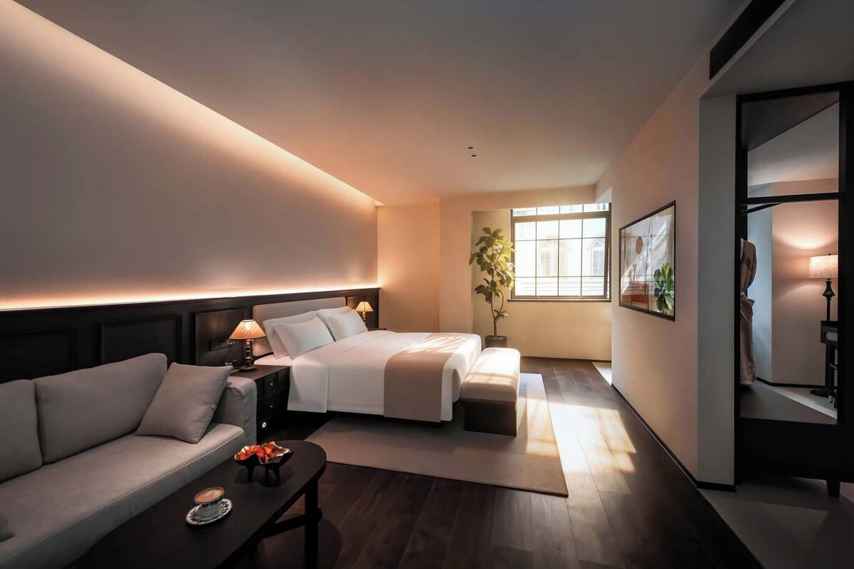 A modern hotel room with a bed, sofa, bedside table, lamp, and a large window.