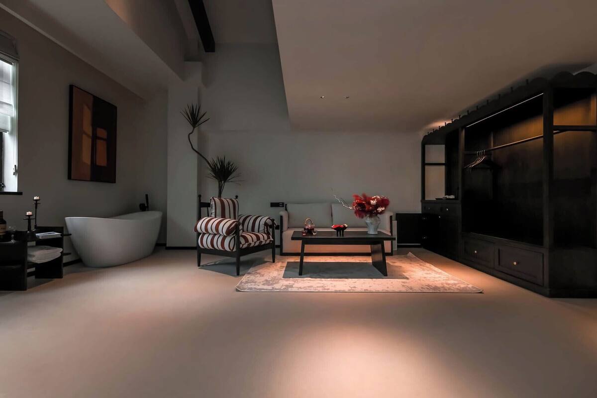 A modern living room with a large white bathtub, a striped armchair, a sofa, a coffee table with red flowers, and a dark entertainment unit.