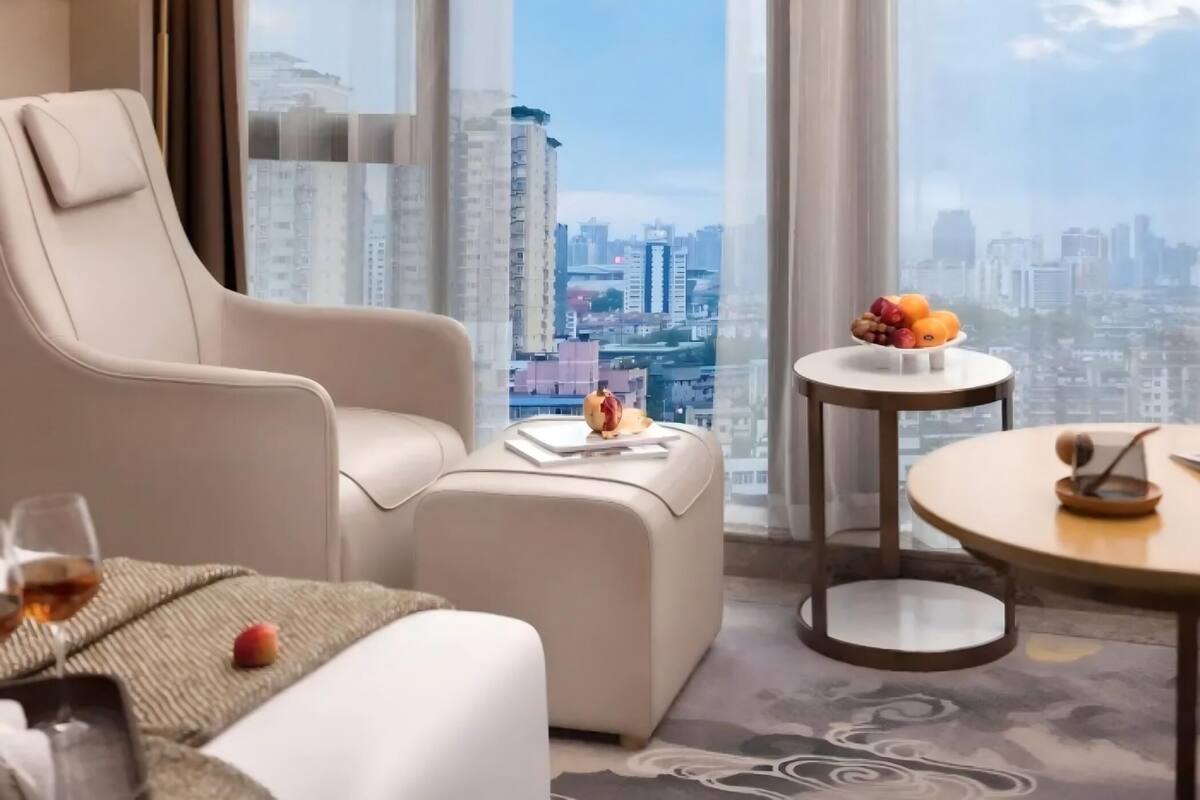 A modern hotel room with a large window offering a city view, a comfortable sofa, a small round table with fruit, and a lamp.