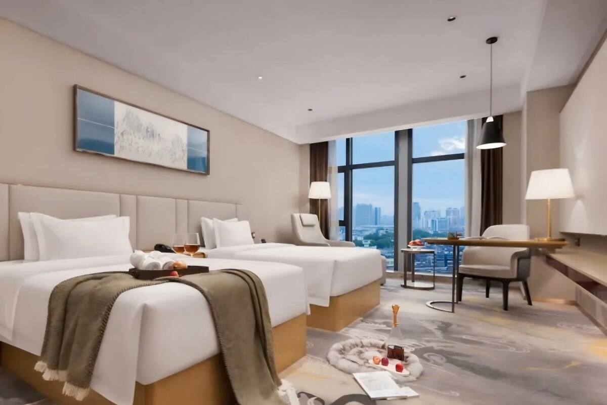 A hotel room with two beds, a desk, a chair, a large window with a city view, and a painting on the wall.