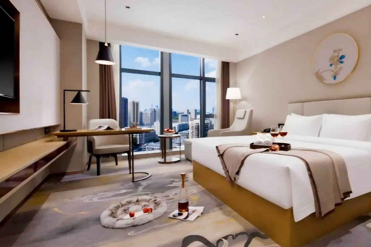 A modern hotel room with a large bed, a desk, a chair, a TV, and a view of the cityscape.