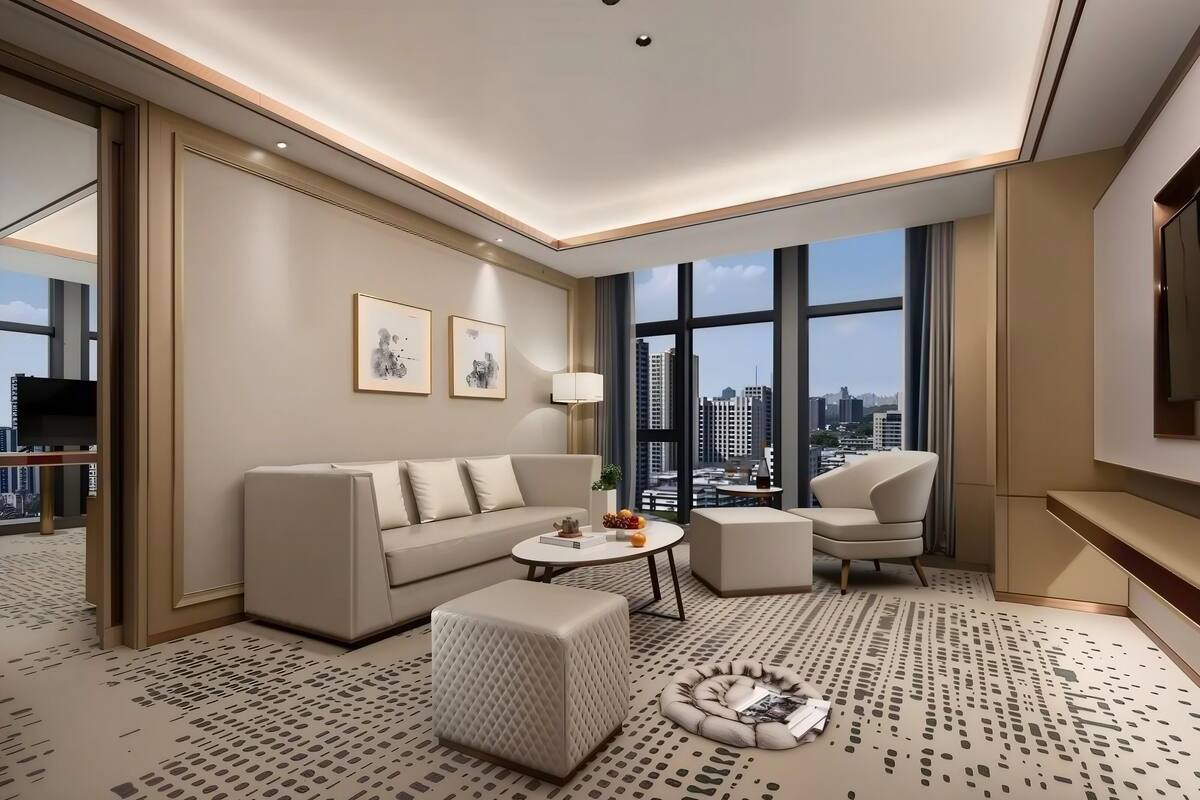 A modern hotel room with a sofa, armchair, ottoman, round coffee table, and a view of the city through large windows.