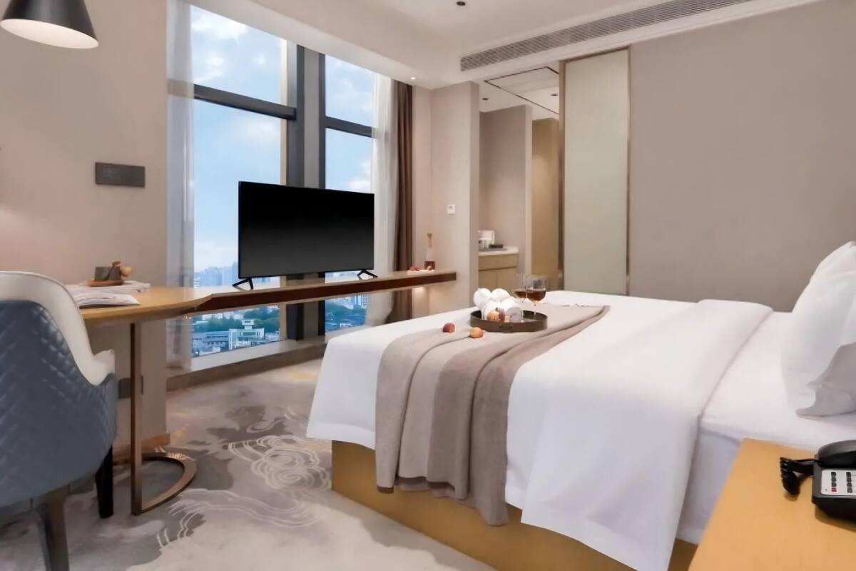 A hotel room with a large window, a desk with a flat-screen TV, a bed with a beige blanket, and a bedside table with a phone and a tray with fruits.
