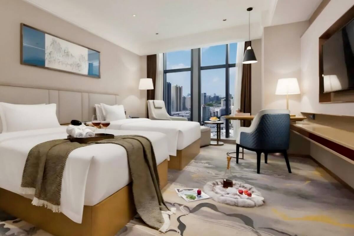 A hotel room with two beds, a desk, a chair, a TV, and a city view.