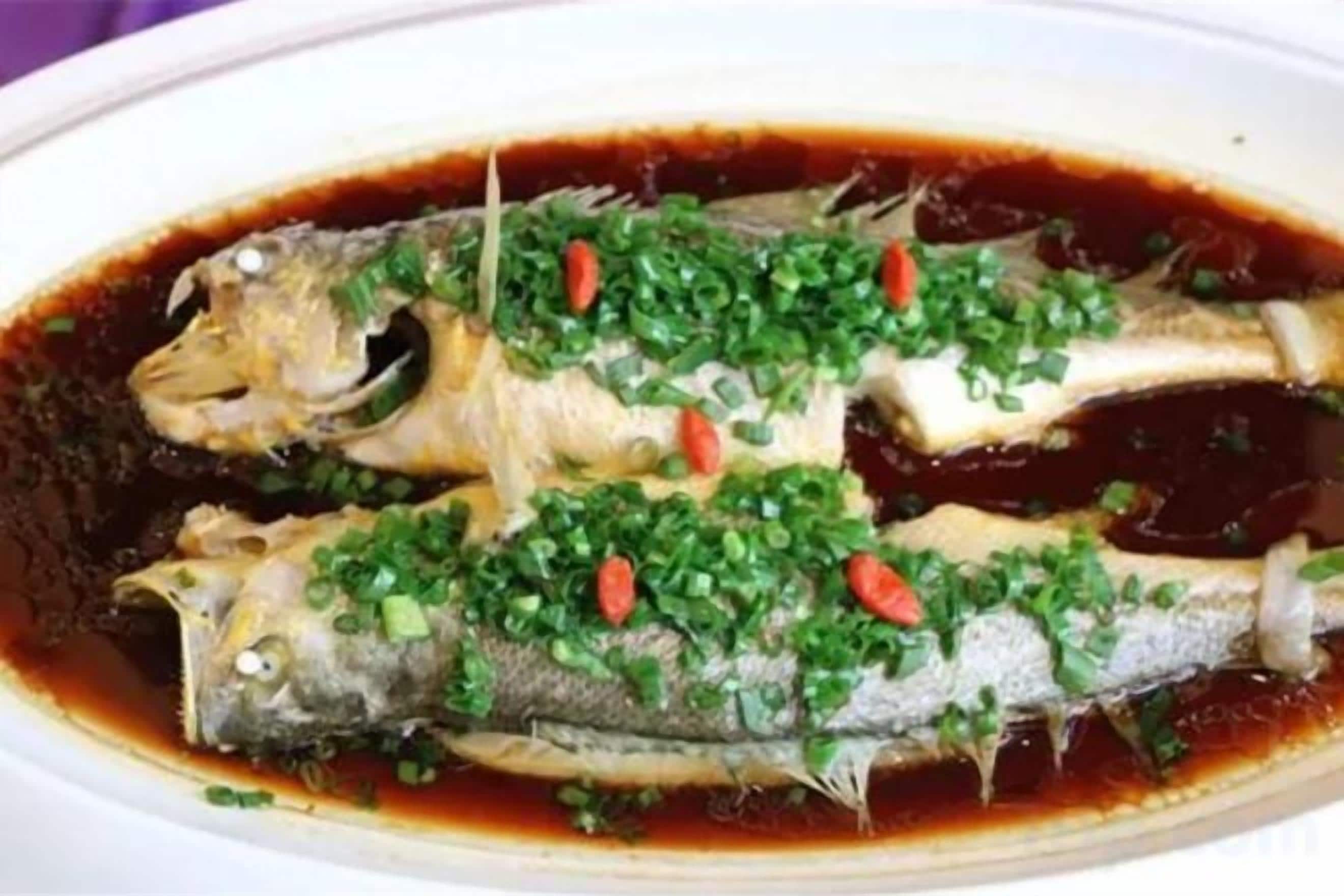 A dish of fish in a dark sauce, garnished with green herbs and red chili slices.