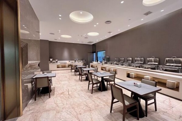 Un vestíbul d'hotel modern amb sòls de marbre, una llarga recepció amb diverses màquines de cafè, i diverses taules i cadires disposades per dinar.