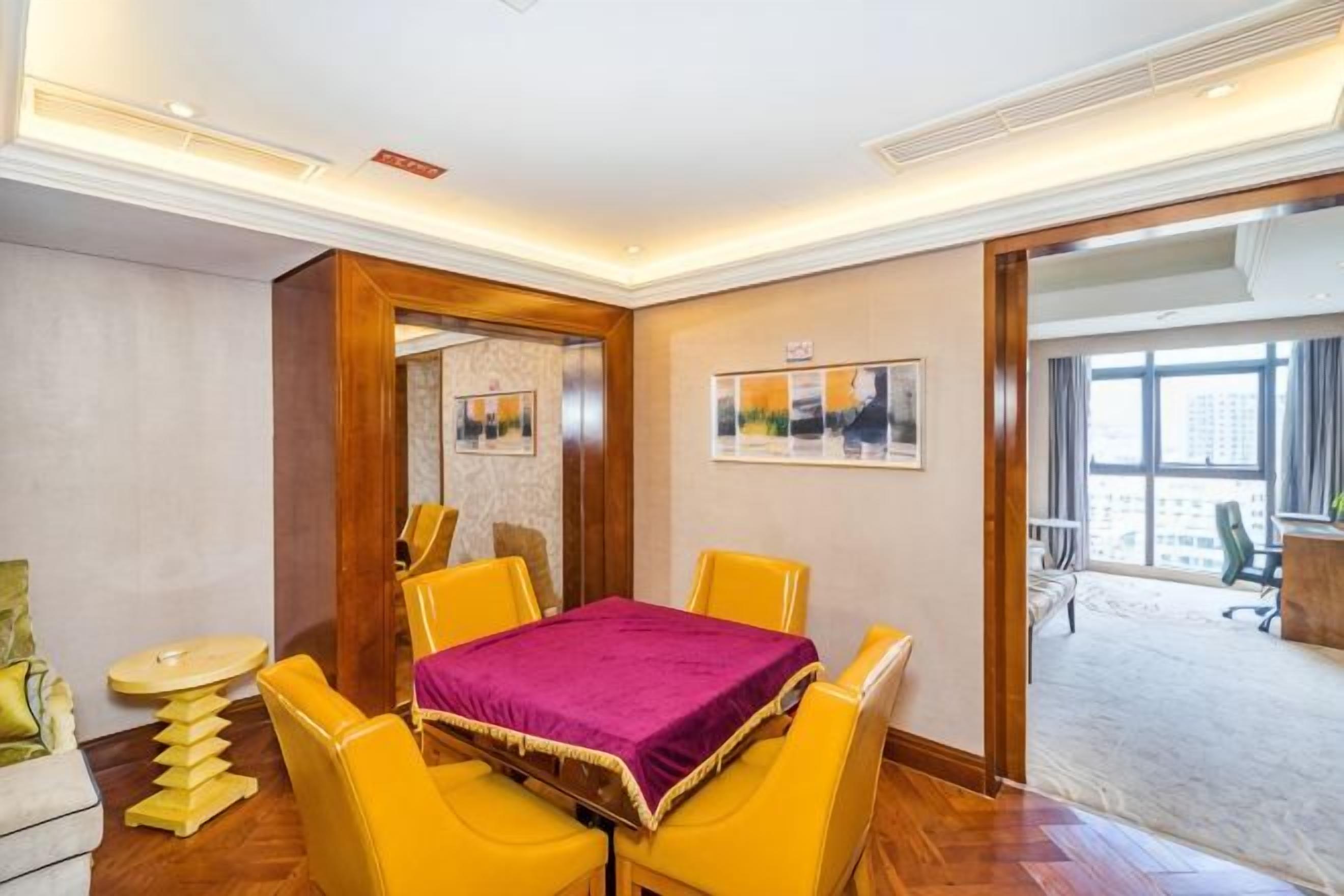 Habitación de hotel con una mesa de comedor, sillas amarillas, un mantel morado y vistas de la ciudad a través de amplios ventanales.