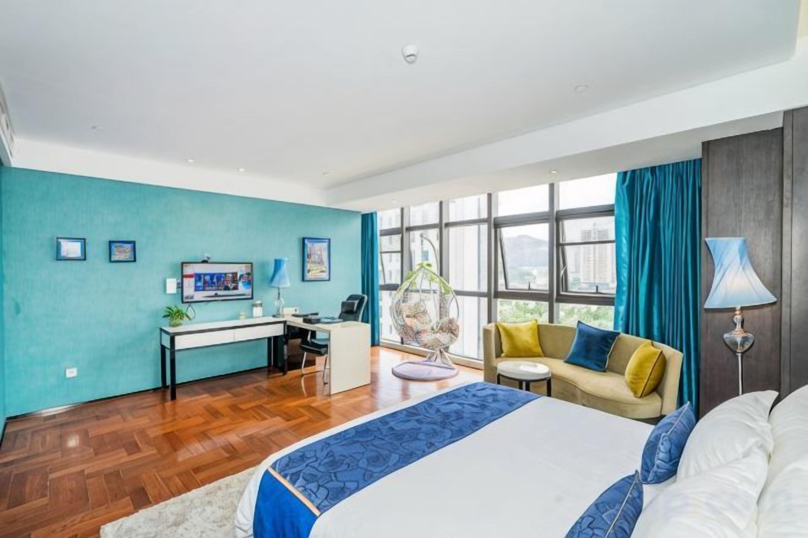 A modern hotel room with a large bed, a desk, a sofa, and a view of the city through large windows.