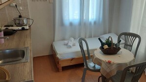 Ein kleines Zimmer mit einem Bett, einem Tisch mit einem Korb, einem Stuhl, einem Waschbecken und einem Fenster mit Vorhängen.