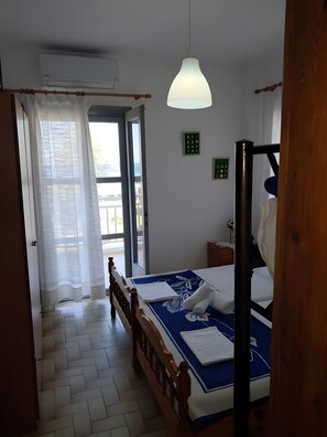 A bedroom with a bed, a wooden headboard, a hanging light fixture, a window with curtains, and a small bedside table. - Apartment 'Marina's Houses By The Sea - Apt 6' with Sea View, Wi-Fi and Air Conditioning (Kassandra)