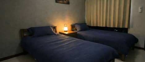 Habitación de hotel con dos camas individuales, una mesita de noche con lámpara, un cuadro en la pared y una ventana con cortinas.