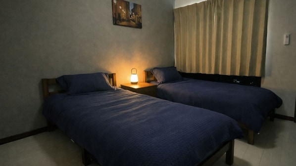 Habitación de hotel con dos camas individuales, una mesita de noche con lámpara, un cuadro en la pared y una ventana con cortinas.