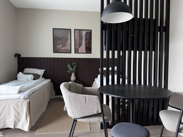 Una habitación de hotel moderna con una cama, dos sillones, una mesa redonda, una planta en maceta y dos cuadros en la pared.