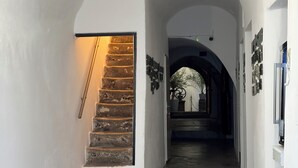 En gang med en stengulvstrappe, hvælvede lofter og udsigt til et rum med et stort vindue og en plante.