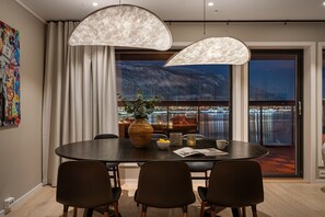 A dining area with a round table, four chairs, two pendant lights, a vase with flowers, a bowl, a candle, and a window with a city view. - Eight floor Premium Waterfront Penthouse (Tromsø)