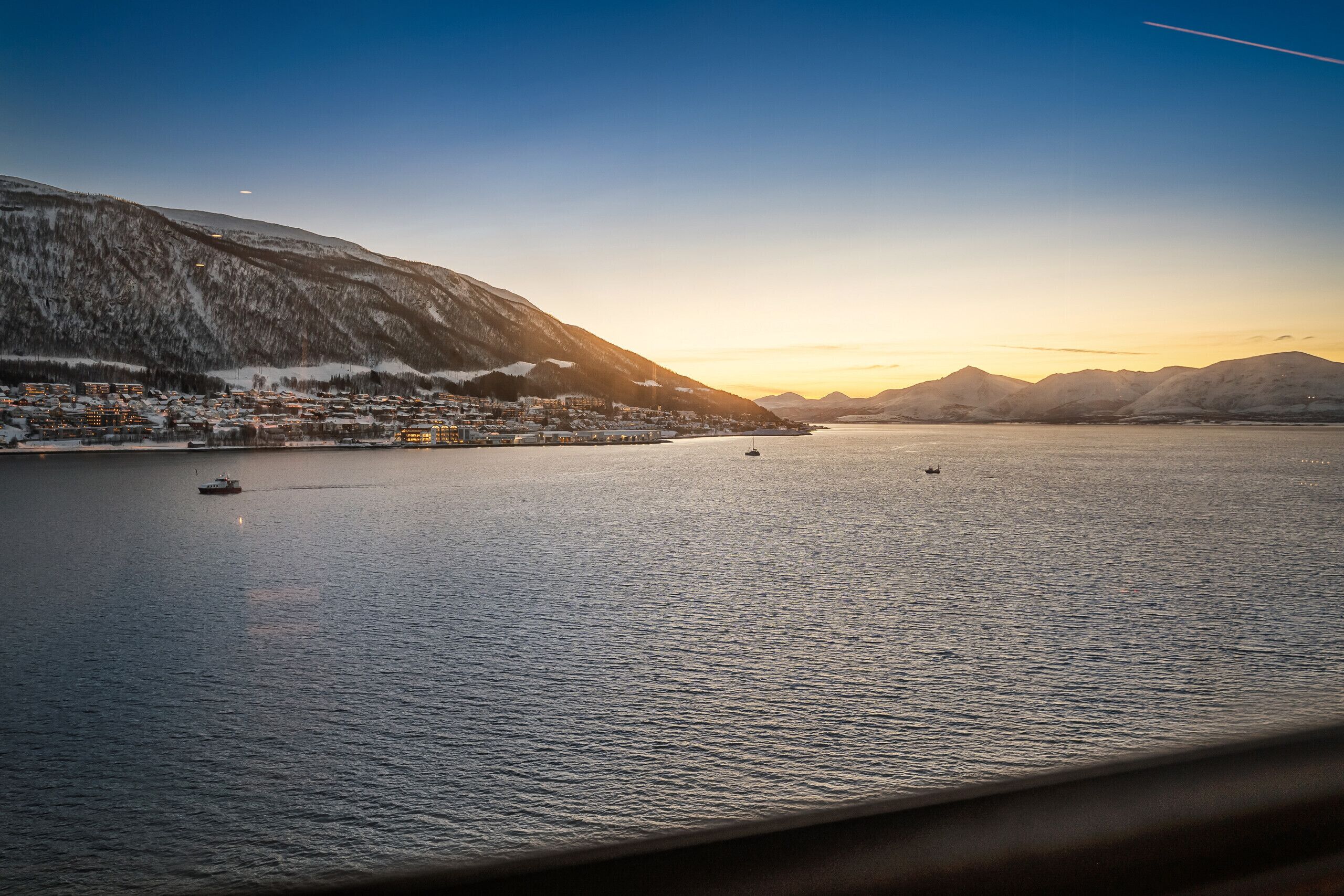 A coastal town with a calm sea, boats, and snow-covered mountains.