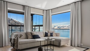 Una sala de estar moderna con un sofá, dos mesas auxiliares, una alfombra y ventanas amplias con vistas a una montaña y un lago.