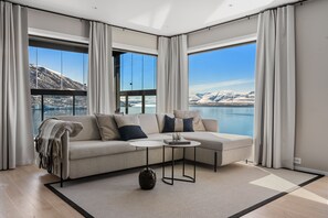 Ein modernes Wohnzimmer mit einer Couch, zwei Beistelltischen, einem Teppich und großen Fenstern mit Blick auf Berge und einen See.