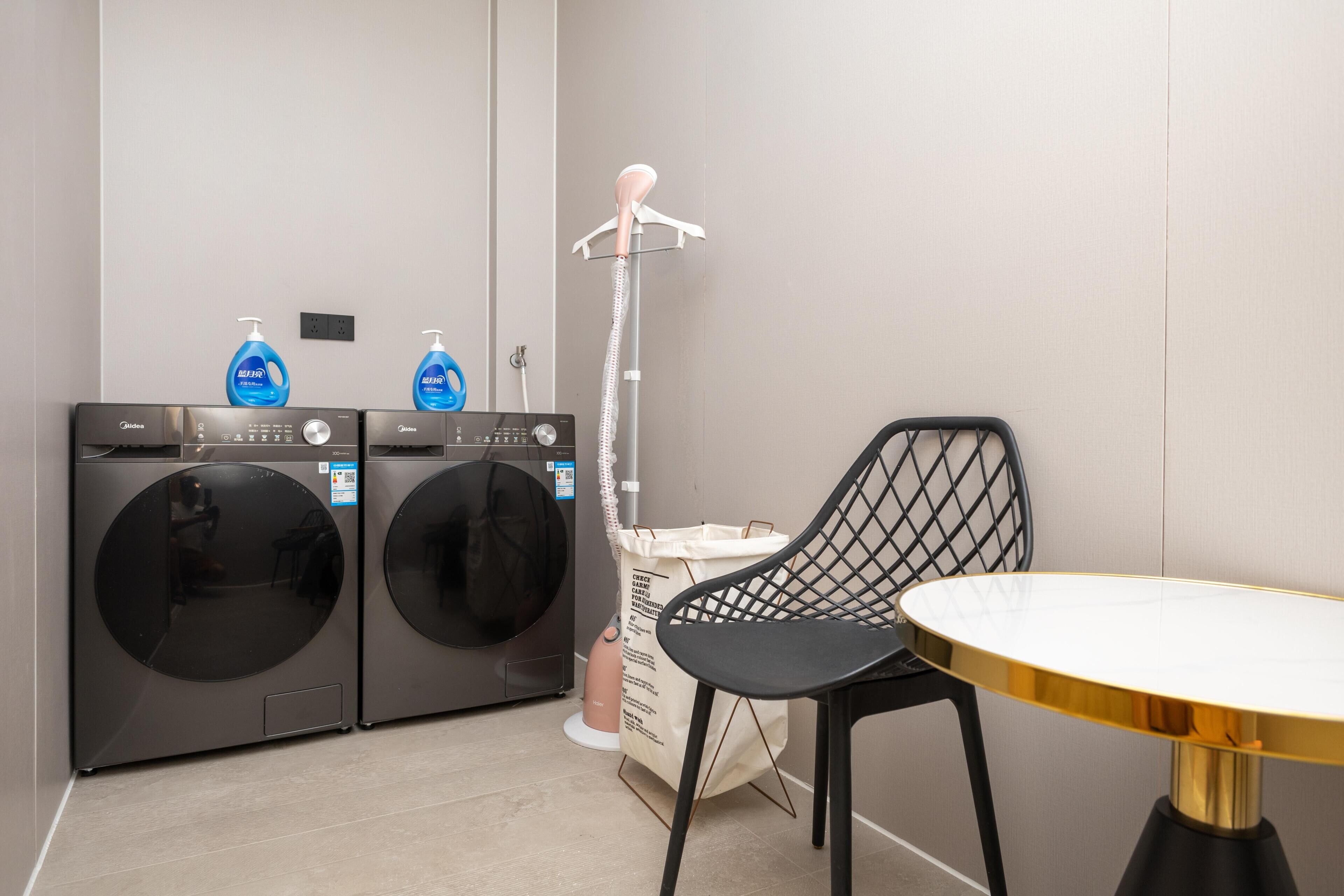 A laundry area with two front-loading washing machines, a drying machine, a hanging rack, a table, and a chair.