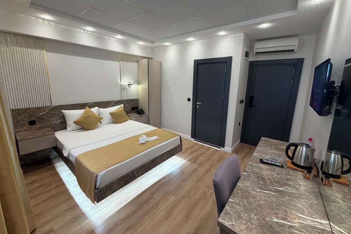 Habitación de hotel con cama, mesita de noche, lámpara, armario, escritorio, silla y cafetera.