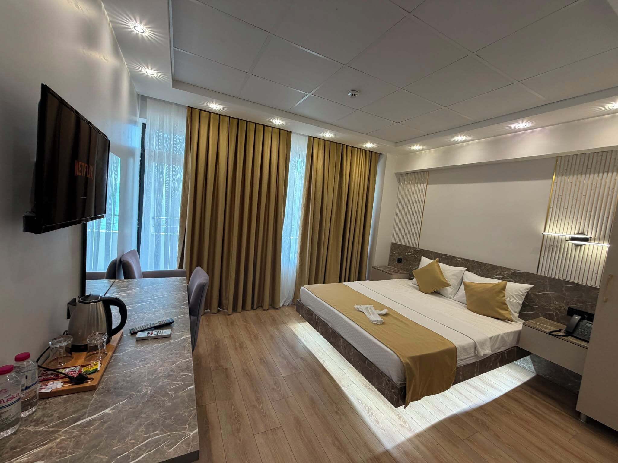 Quarto de hotel com cama, escrivaninha com chaleira, televisão e janela com cortinas.