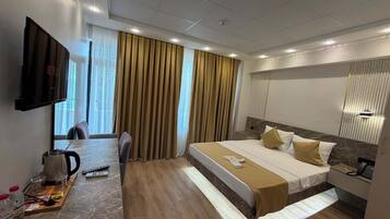 Quarto de hotel com cama, escrivaninha com chaleira, televisão e janela com cortinas.