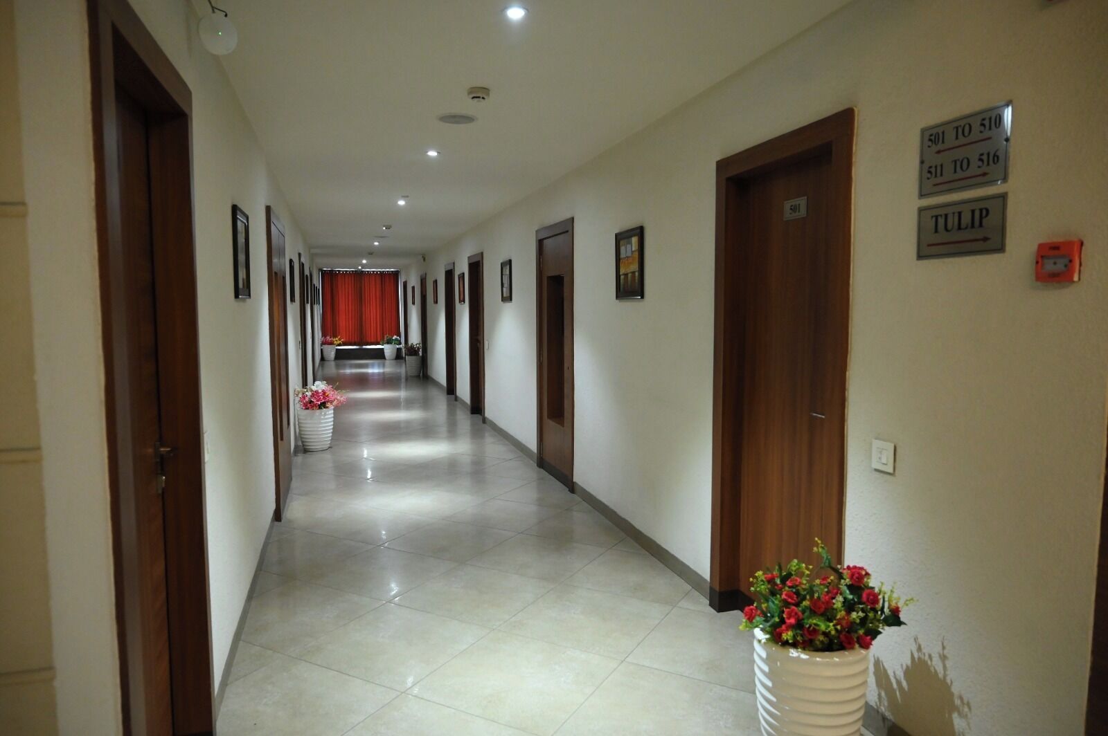 Um corredor de hotel com portas numeradas de 501 a 516, uma cortina vermelha ao fundo e vasos de flores ao longo das paredes.